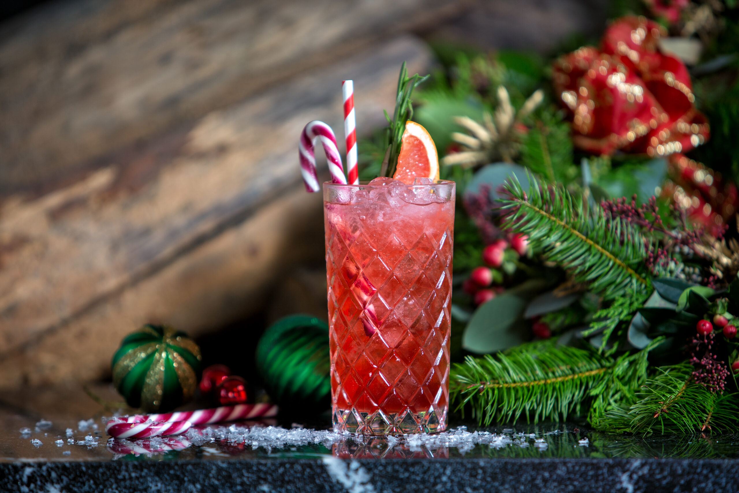 festive red coloured drink with candy cane and orange garnish. Christmas foliage in the background on a wood table with some dusting on top